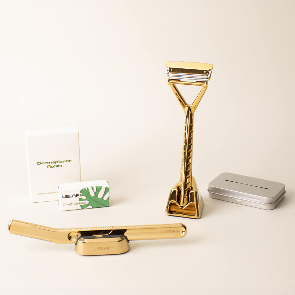 The gold full face and body kit displayed with the leaf razor, dermaplaner, a stand and blades, for each, and the recycling tin